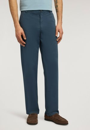 Mand iført navyblå bukser med lige ben, lyseblå skjorte, brune loafers og en kinesisk tegn-tatovering på venstre underarm.