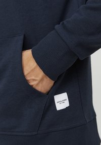 Námořnická modrá mikina s žebrovanými manžetami a kapsou. Na dolní levé straně je našitá látková nálepka s nápisem "JACK&JONES EST.1990."