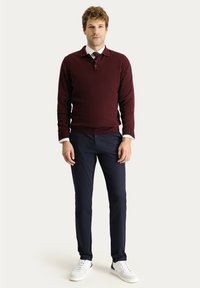 Bordeauxfarbener Strickpullover mit Kragen, über einem weißen Hemd und einer Krawatte getragen. Dunkelblaue Hose und weiße Sneakers runden das Outfit ab.