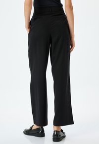 Pantalones de pierna ancha negros fabricados en un tejido suave, con una cinturilla de altura media y bolsillos laterales. Combinados con mocasines negros brillantes.