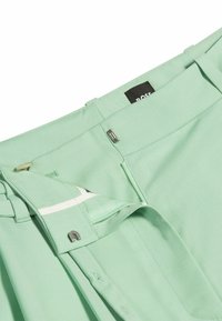 Mintgröna byxor med en slät tygstruktur, utrustade med dragkedja och knappstängning, inre vit tejp och en svart etikett.