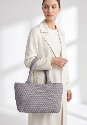 Femme en manteau blanc tenant un grand sac fourre-tout gris tissé avec deux anses sur un fond lumineux et minimaliste.