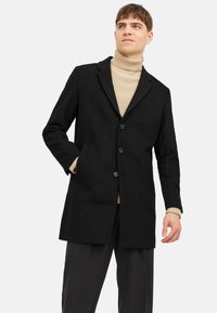 Schwarzer Wollmischungsmantel mit strukturierter Form, einreihigem Design, Reverskragen und Seitentaschen, getragen über einem beigefarbenen Rollkragenpullover.
