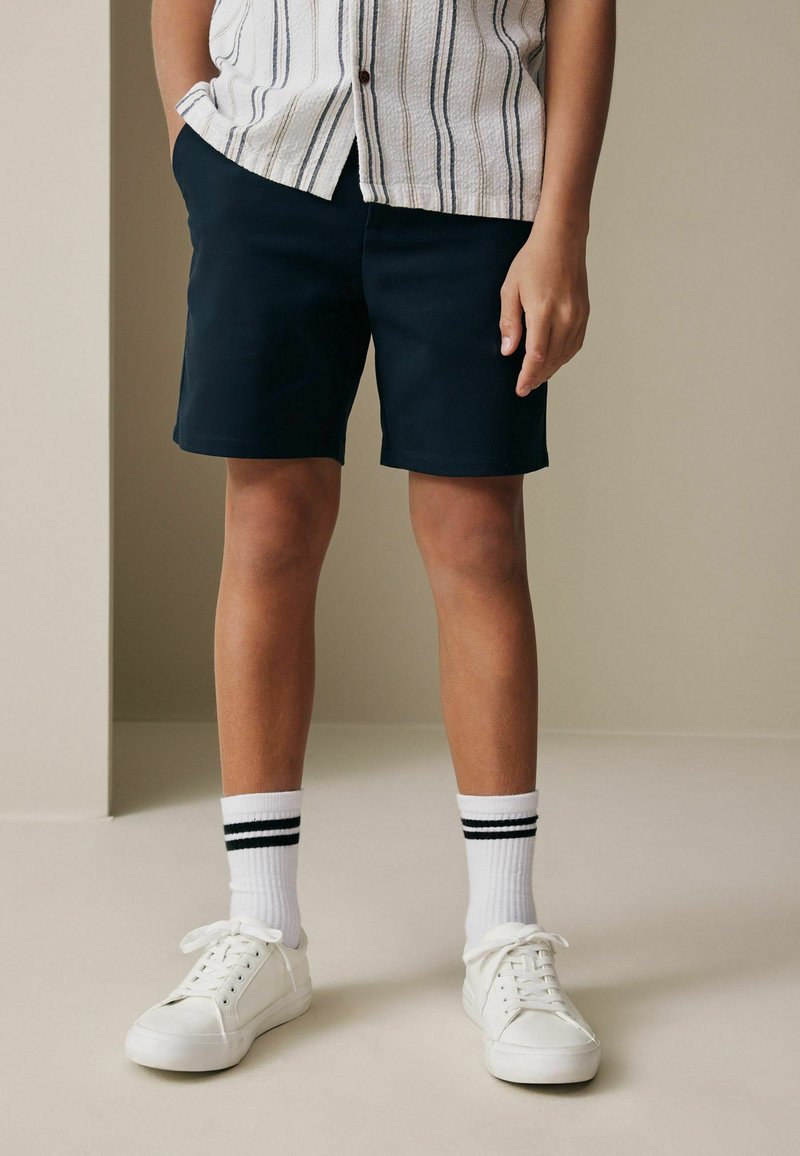 Marineblaue Shorts mit maßgeschneiderter Passform, kombiniert mit weißen Sneakers und kniehohen Socken mit schwarzen Streifen. Glatter Stoff mit klaren Linien.