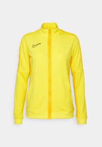 Nike keltainen vetoketjullinen takki korkealla kauluksella, musta logo vasemmassa rinnassa, pitkät hihat ja sivutaskut, valmistettu sileästä, joustavasta kankaasta.