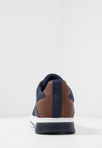 Navy blue sneaker featuring a textured mesh upper, brown leather accents, extended heel tab, and white rubber sole with "bugatti" branding.