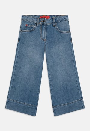 Jean en denim bleu à jambes larges cropped avec poches avant, passants de ceinture, fermeture par bouton et une étiquette de marque rouge à l'intérieur de la ceinture.