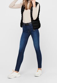 Černá bundová vesta přes béžové tričko s dlouhým rukávem, v kombinaci s tmavomodrými skinny jeans a bílými teniskami s oranžovými detaily, ve vzpřímené pozici.