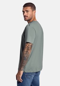 Camiseta gris de manga corta, cuello redondo, corte relajado, con un acabado texturizado. Presenta tatuajes visibles en el brazo. Se lleva con unos vaqueros azules.