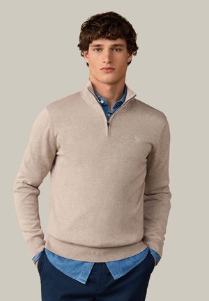 Jonge man met krullend haar, gekleed in een beige trui met een kwart rits over een blauwe overhemd met knopen en donkere broek, staand met zijn handen in zijn zakken.