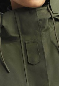 Veste imperméable vert olive avec un col haut, poche avant et capuche avec cordon de serrage. Texture lisse et détails de couture subtils.