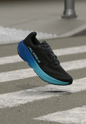 Zwart-blauwe hardloopschoen met "ALTRA" branding die boven een zebrapad op een asfaltweg met kleine grinddeeltjes zweeft.