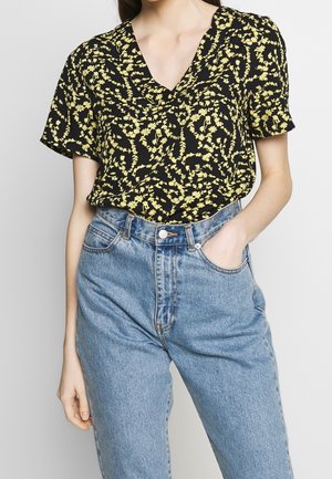Blusa nera a maniche corte con un motivo floreale giallo e scollatura a V, abbinata a jeans in denim azzurri a vita alta.