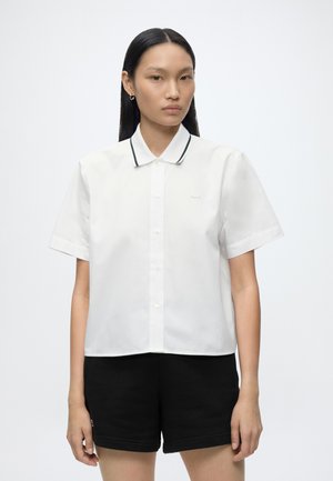 Vrouw met lang zwart haar draagt een wit overhemd met korte mouwen en knopen, met een kraag afgezet met zwart, en zwarte korte broek, staand tegen een eenvoudige achtergrond.