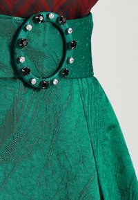 Gonna di tessuto verde con una finitura testurizzata, caratterizzata da una cintura larga adornata con una fibbia circolare, accentuata da gemme nere e trasparenti.