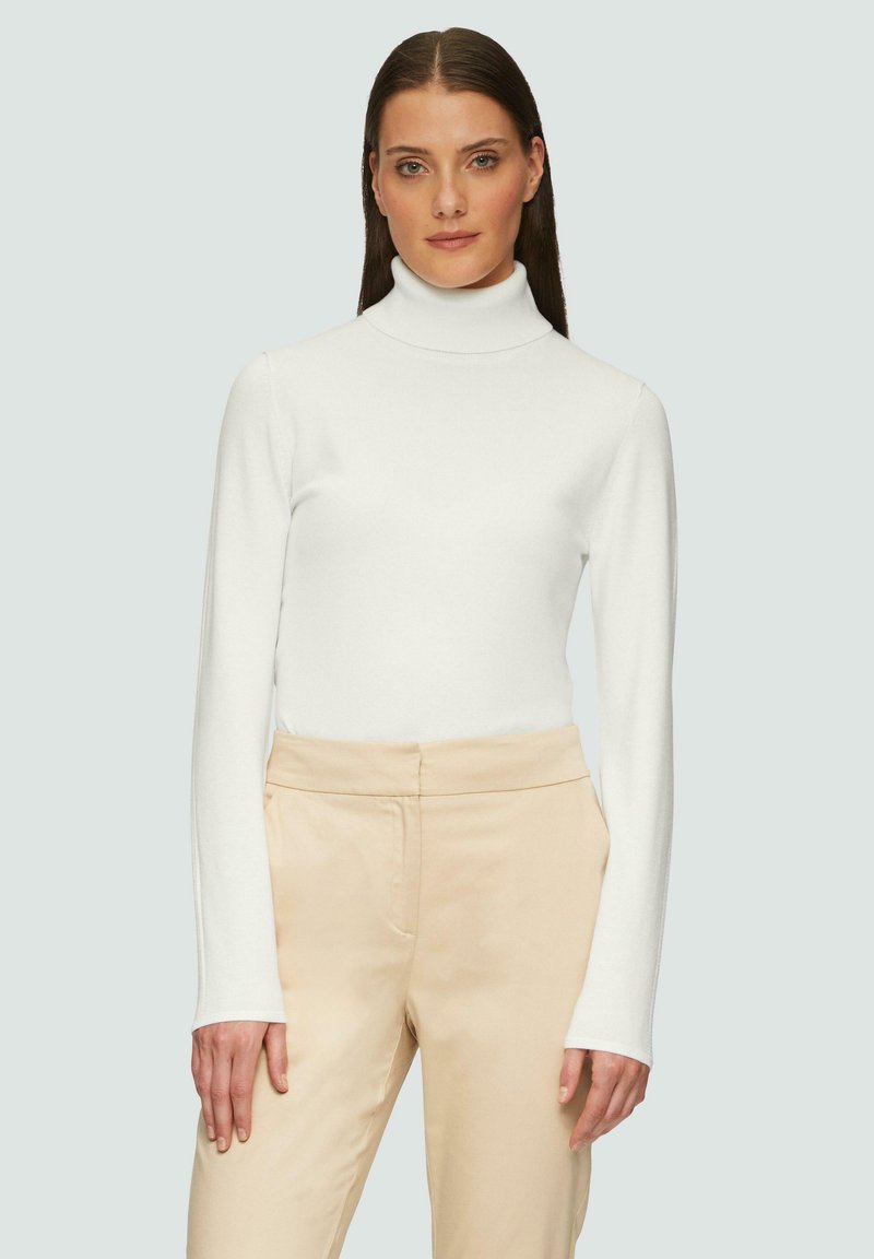 Haut à col roulé blanc avec des manches longues, texture lisse, design ajusté. Associé à un pantalon taille haute beige, style minimaliste.