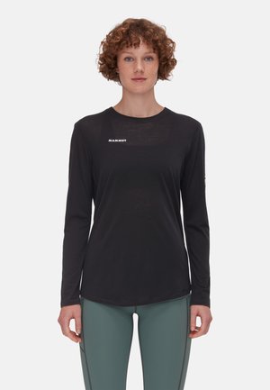 Langärmliges schwarzes Shirt aus leichtem Stoff mit rundem Halsausschnitt und Logo auf der Brust; kombiniert mit grünen, eng anliegenden Hosen.
