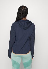 Donna con lunghi capelli intrecciati che indossa una giacca con cappuccio blu navy con rifiniture arancioni e leggings chiari a fantasia verde, rivolta di spalle contro uno sfondo semplice.