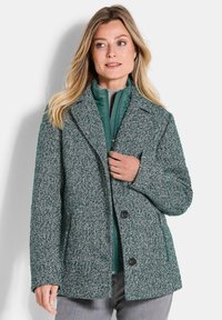 Femme portant un manteau en laine textur�é vert superposé et une veste zippée matelassée, se tenant devant un fond clair.