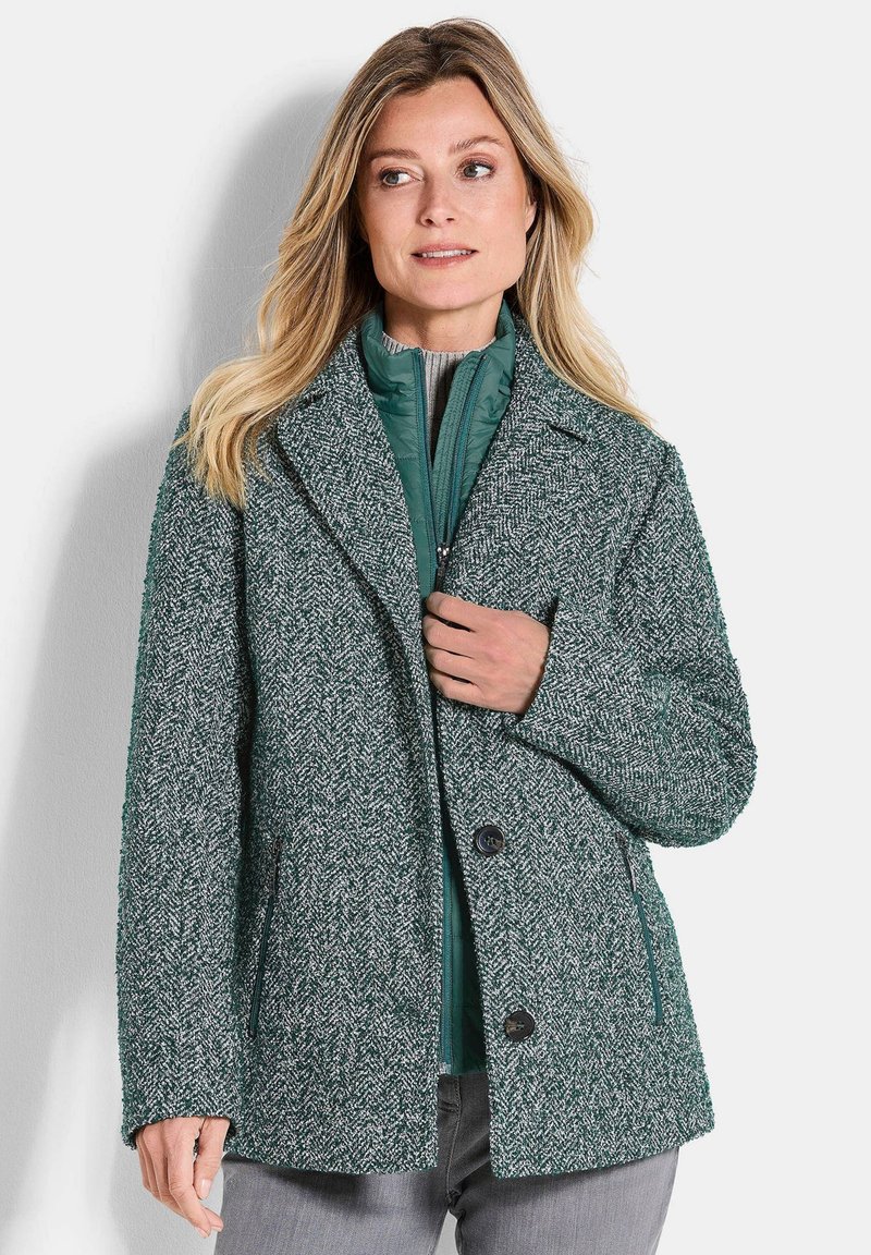 Frau in einem gestuften, grünen, strukturierten Wollmantel und einer gepolsterten Zip-Jacke, die vor einem schlichten, hellen Hintergrund steht.