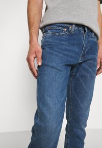 Mörkblå denimjeans med rak skärning. Funktioner inkluderar en dragkedja, kopparnitar och subtilt slitning på tyget.