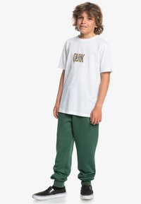 Camiseta blanca de algodón con gráfico "QUIK", combinada con pantalones de chándal verdes y zapatos negros sin cordones. Ajuste casual y textura suave.