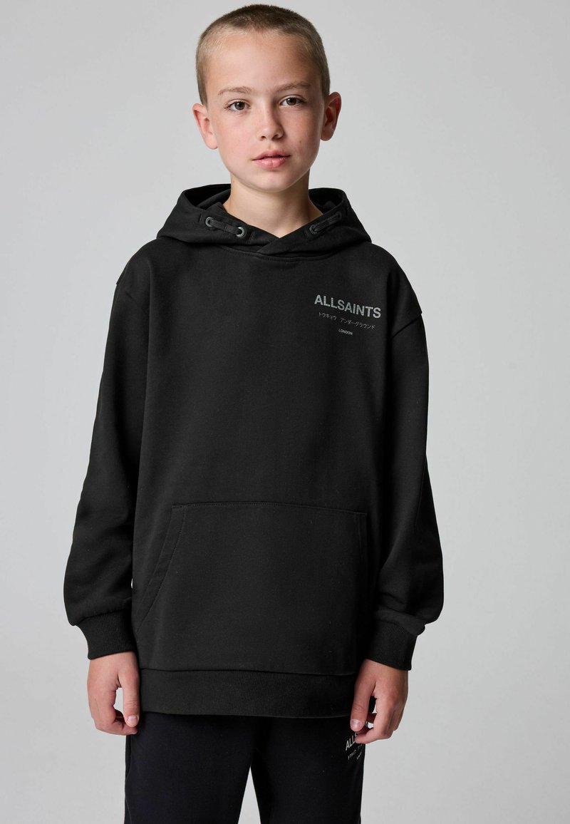 Fant je oblečen v črno kapuco AllSaints in usklajene hlače, stoji ob ozadju v enotni sivi barvi, obrnil se je naprej z nevtralnim izrazom na obrazu.