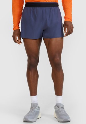 Mężczyzna ubrany w granatowe sportowe krótkie spodenki, szare buty do biegania z białymi skarpetkami oraz pomarańczową bluzę z długim rękawem, stojący na jednolitym tle.