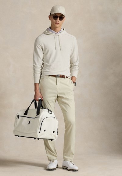 Λευκή τσάντα duffle με μαύρες λεπτομέρειες, φορεμένη με ανοιχτό μπεζ φούτερ με κουκούλα και παντελόνια. Τα αξεσουάρ περιλαμβάνουν καπέλο και γυαλιά ηλίου.
