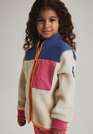 Enfant aux cheveux bouclés portant une veste en polaire crème avec un empiècement bleu, une poche poitrine rose et une fermeture éclair orange, debout devant un fond clair.