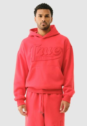 Man wearing red "True Religion" hoodie and matching red sweatpants against plain light background.