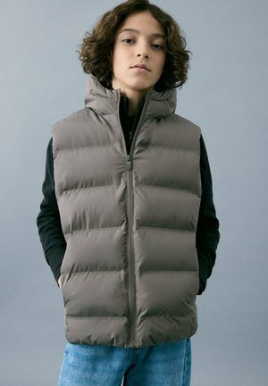 Veste matelassée grise sans manches avec col montant, fermeture éclair à l'avant et motif matelassé horizontal. Portée sur un haut à manches longues noir et un jean en denim bleu.