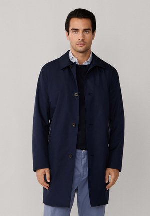 Man wearing a navy blue single-breasted coat over a black sweater and striped shirt, paired with light blue trousers, standing against a plain background.