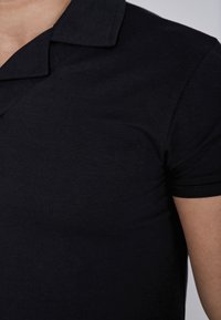 Melnā polo krekls ar apkakli, īsām piedurknēm un gludu, vieglu audumu. Iezīmē klasisku, pieguļošu dizainu.