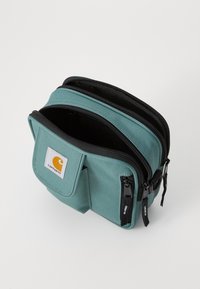 Geantă din material teal cu două fermoare, un buzunar frontal și detalii negre. Prezintă un patch cu logo pe partea din față. Design compact și rectangular.
