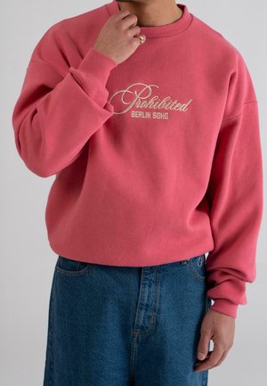 Personne portant un sweat-shirt rose avec le texte brodé "Prohibited Berlin Soho", associé à un jean bleu, main près du visage montrant une bague.