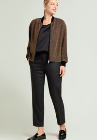 Glinsterend bronzen bomberjack met zwarte accenten, gecombineerd met op maat gemaakte zwarte pantalon met krijtstreep en zwarte loafers, op een lichte achtergrond.