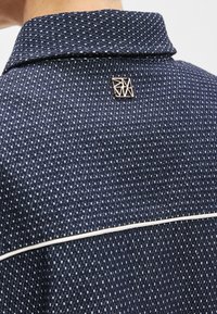 Μπλε πουκάμισο με γιακά, κατασκευασμένο από υφασμά με υφή και διακριτικό σχέδιο. Περιλαμβάνει μια μεταλλική γεωμετρική καρφίτσα και λευκή λεπτομέρεια με ρίγα.