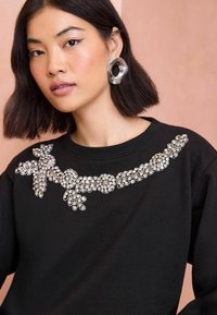 Crni dukserica s dekorativnim ukrasima od rhinestona preko grudi, s cvjetnim i kružnim dizajnima. Okrugli srebrni naušnica je vidljiv.