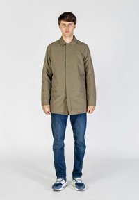 Cappotto khaki a maniche lunghe con colletto, chiusura a bottoni, due tasche laterali, abbinato a jeans blu e sneakers blu, in piedi davanti a uno sfondo neutro.