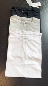 Tre foldede t-shirts i sort, grå og hvid. Rund hals design, bomuldsstof, og mærket synligt med størrelsesmærkater. Glat tekstur.