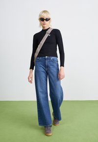 Svart langermet topp, høytlivde blå vide jeans, grå sneakers og katteøy solbriller. Modell går på et grønt gulv.