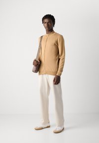 Man in een beige zip-up sweater, off-white broek en witte sneakers, met een grijze schoudertas, staande tegen een effen achtergrond.