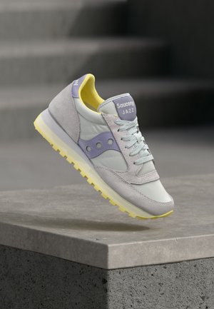 Világosszürke és levendulalila Saucony Jazz tornacipő sárga talppal és belső béléssel, levegőben, egy beton párkány fölött elhelyezve.