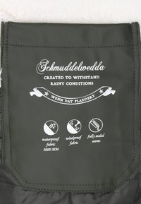 Tumši zaļš ūdensnecaurlaidīgs auduma marķējums ar baltu tekstu, kas apraksta īpašības: ūdensnecaurlaidīgs (3000 mm), vēja necaurlaidīgs un pilnībā pielīmētas šuves.