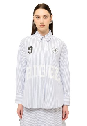 Giovane donna con capelli lisci e scuri che indossa una camicia azzurro chiaro a righe sottili con colletto, numero 9 e toppe con logo, in piedi davanti a uno sfondo bianco.