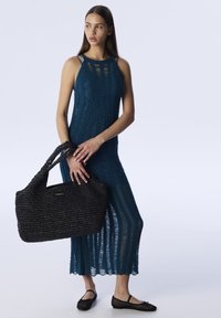 Teal pletené šaty s vertikálními pruhy, doplněné černou tkanou taškou s nápisem "TWIST". Černé slip-on boty dotvářejí vzhled.