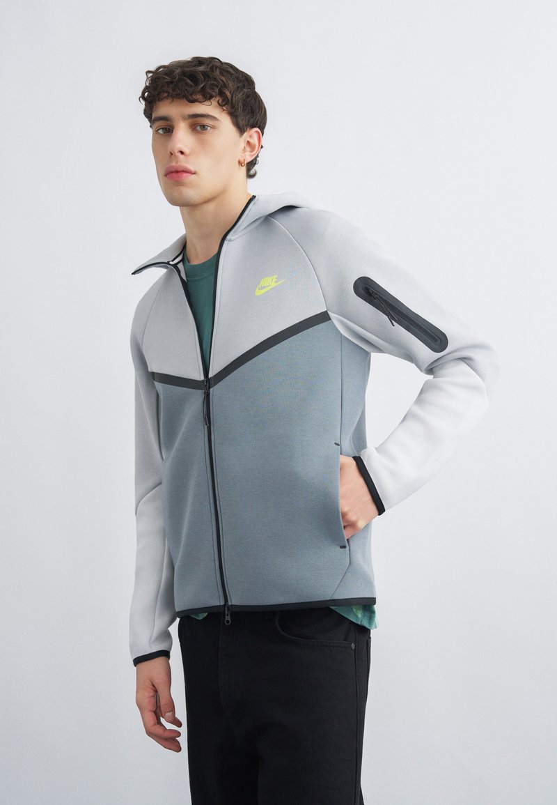 Junger Mann, der eine graue und weiße Nike-Zipjacke mit neonfarbenem Logo trägt, steht mit einer Hand in der Tasche vor einem schlichten hellen Hintergrund.