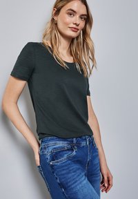 Tmavě zelené tričko s krátkým rukávem a kulatým výstřihem, spárované s modrými džíny. Tkanina vypadá měkká s jemnou texturou.
