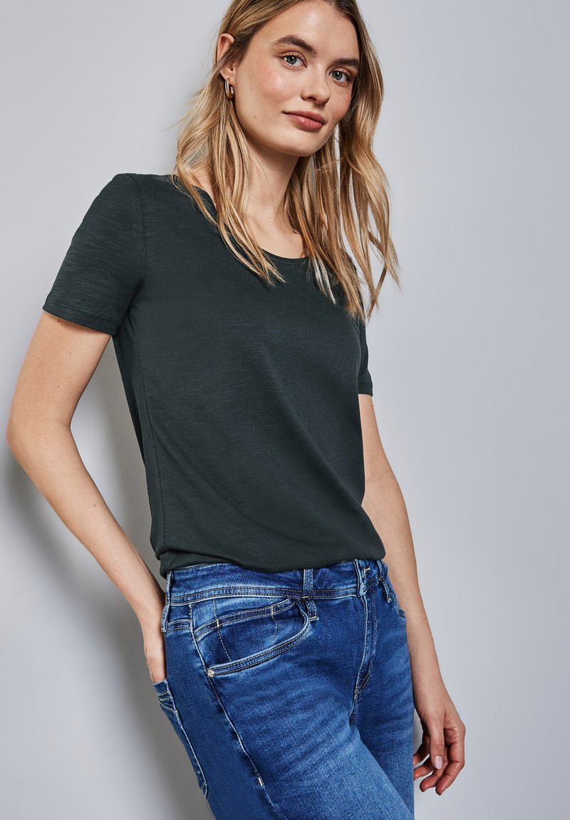 Tmavě zelené tričko s krátkým rukávem a kulatým výstřihem, spárované s modrými džíny. Tkanina vypadá měkká s jemnou texturou.